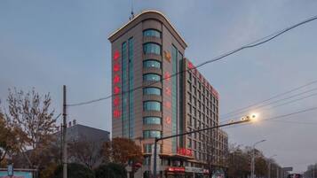 Un hôtel à plusieurs étages, doté d’un design moderne, avec une enseigne verticale bien visible, situé à un carrefour et offrant une vue sur la rue, avec des véhicules et des arbres.