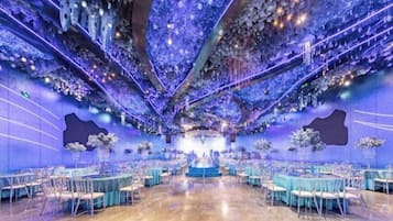 Um salão amplo com palco central, cercado por mesas e cadeiras, e teto decorado com ornamentos pendentes.