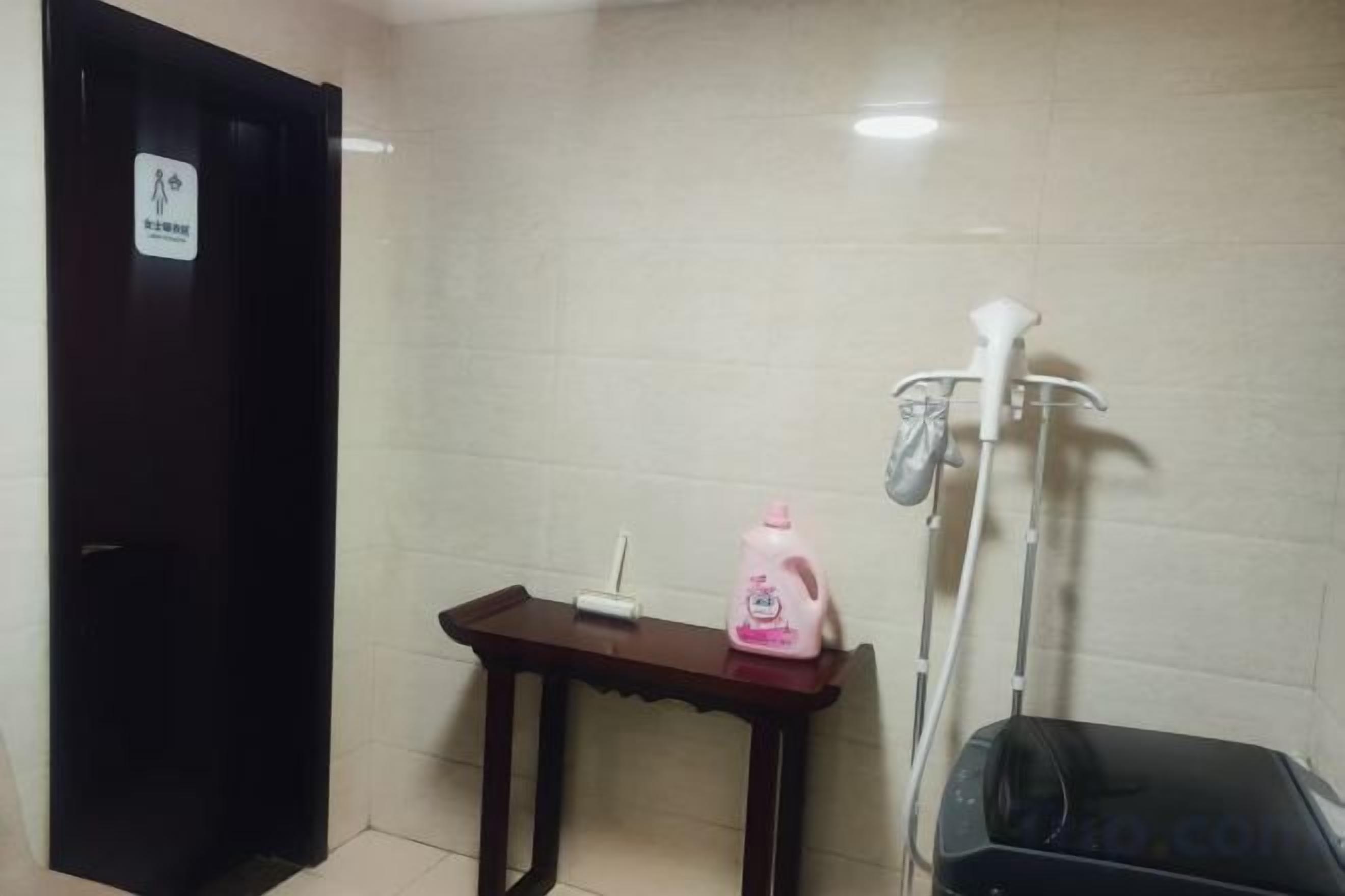 Ein Badezimmer mit einer dunkelholzigen Tür, einem kleinen Tisch mit einer rosa Flasche, einem weißen Duschkopf und einem schwarzen Koffer.