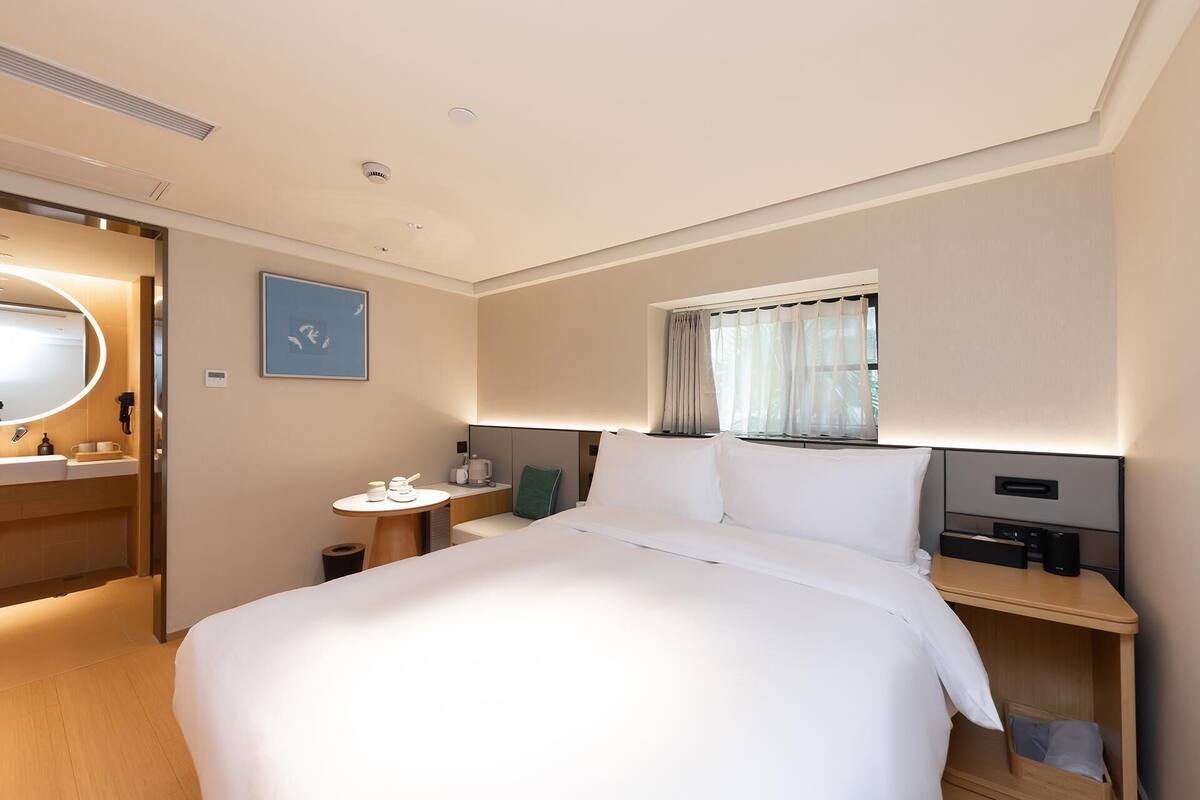 A hotel room with a bed, bedside tables, a desk, a chair, a window with curtains, a mirror, and a framed picture on the wall.