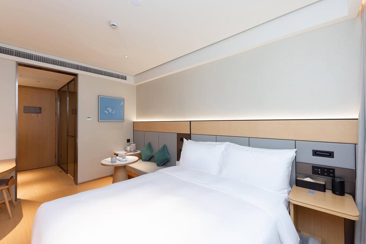 A hotel room with a bed, bedside tables, a nightstand, a desk, a chair, a door, and a wall-mounted light fixture.