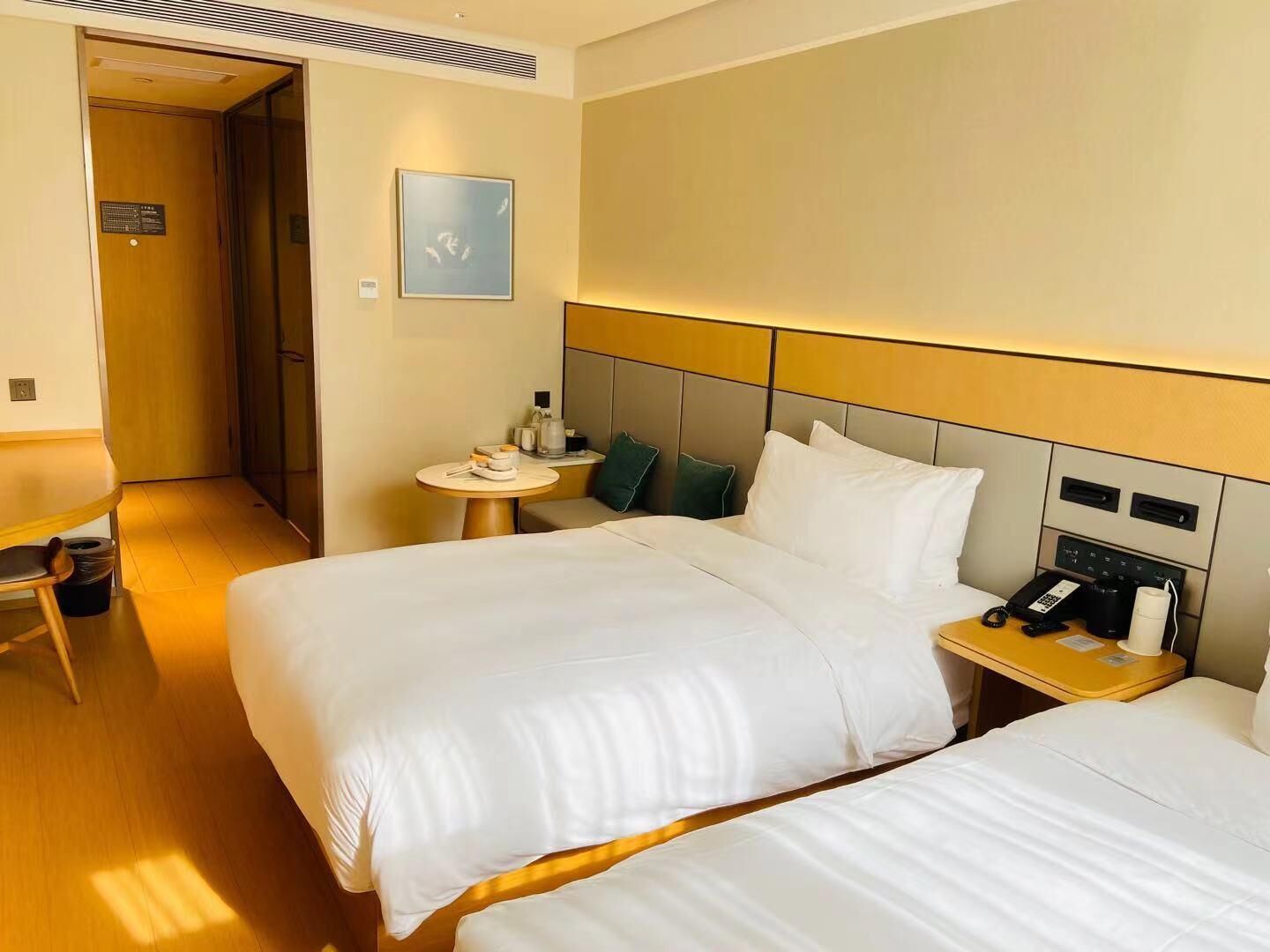 Habitación de hotel con dos camas, una mesita de noche con teléfono y cafetera, una pequeña mesa redonda con lámpara y un cuadro en la pared.
