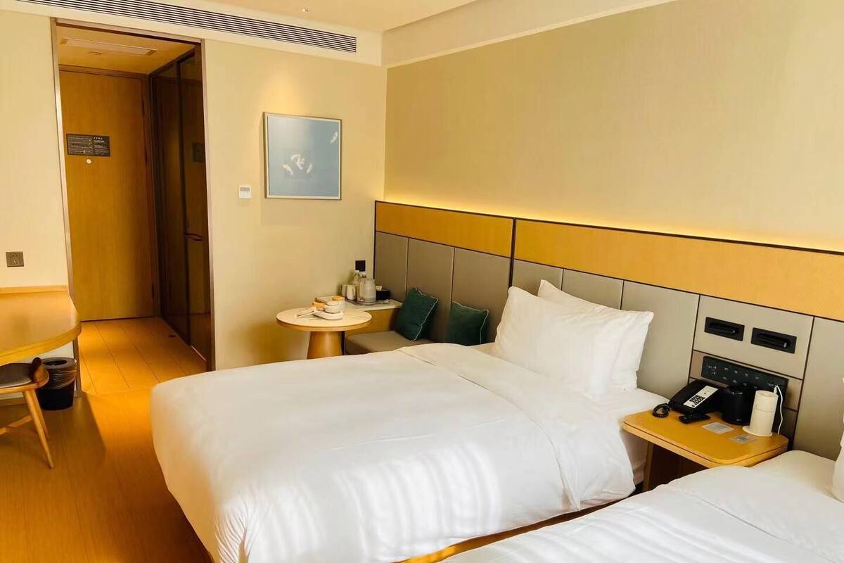 A hotel room with two beds, a bedside table with a phone and a coffee maker, a small round table with a lamp, and a painting on the wall.