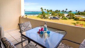 A balcony with a glass table, two chairs, a bottle of liquor, and two glasses. The view includes a beach, palm trees, and a parked car.