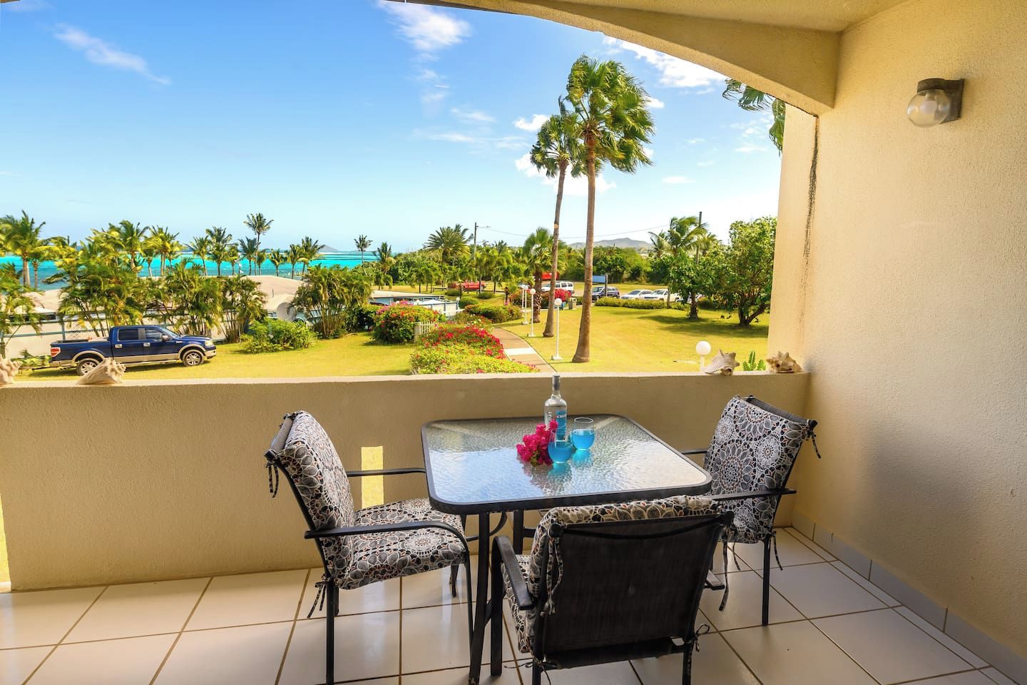 A balcony with a table and chairs, overlooking a parking area and a view of palm trees and a beach.