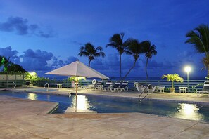 A pool area with lounge chairs and umbrellas, palm trees, and a view of the ocean at dusk. - Comfy condo minutes from downtown & Free Parking (Christiansted)