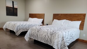Dos camas dobles con ropa de cama blanca y un estampado de ramas de árbol, un cabecero de madera y una obra de arte colgada en la pared que muestra una cerca de alambre de púas.