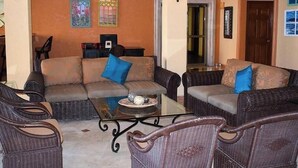 Un salón amplio con mobiliario de mimbre, una mesa de centro de vidrio y una pared decorativa con vidrieras.