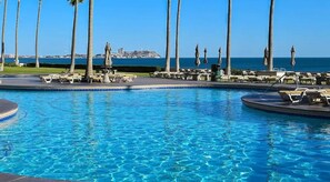 A pool area with lounge chairs and umbrellas, palm trees, and a view of the sea and a distant island. - Beachfront Upper Floor Luxury Condo (Puerto Peñasco)