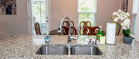 A kitchen with a granite countertop, stainless steel sink, and a double faucet. There are chairs, a table, and a potted orchid.