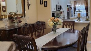 Un comedor con una gran mesa de madera, sillas ornamentadas, una araña y una cocina al fondo.