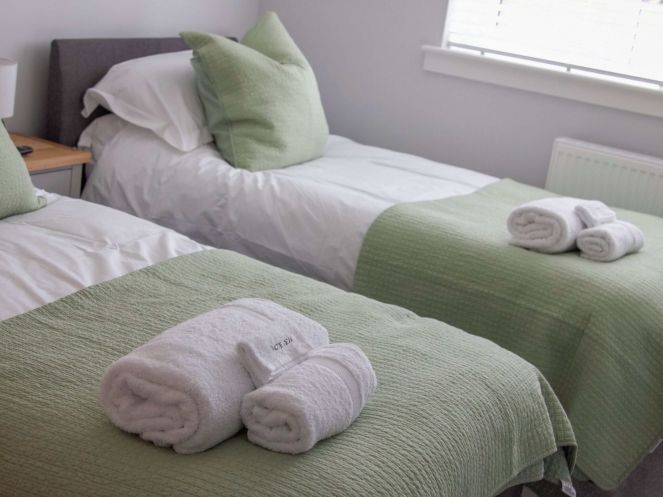Et hotelværelse med to senge, begge med grønne sengetæpper og hvide sengetøj, samt et par sammenrullede håndklæder på sengen i forgrunden.