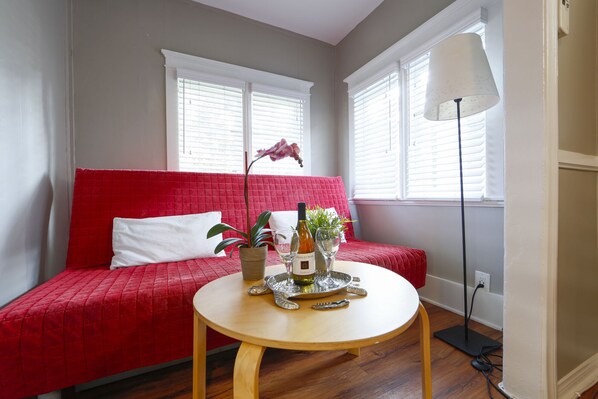 Una habitación con un sofá rojo, una mesa redonda de madera con una botella y vasos, y una lámpara de pie.