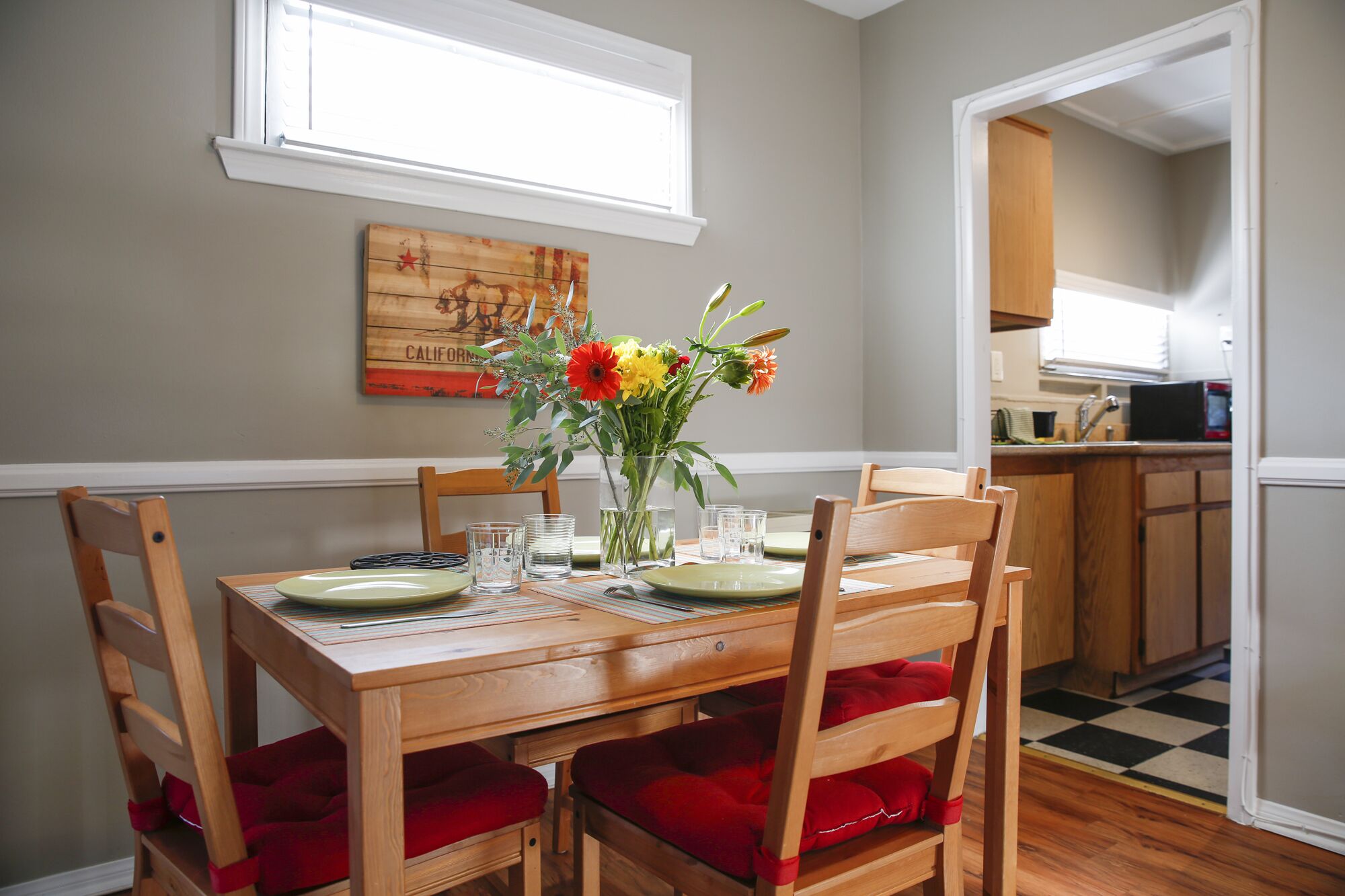 Un comedor con una mesa de madera, sillas con cojines rojos, un florero con flores y un cuadro en la pared.