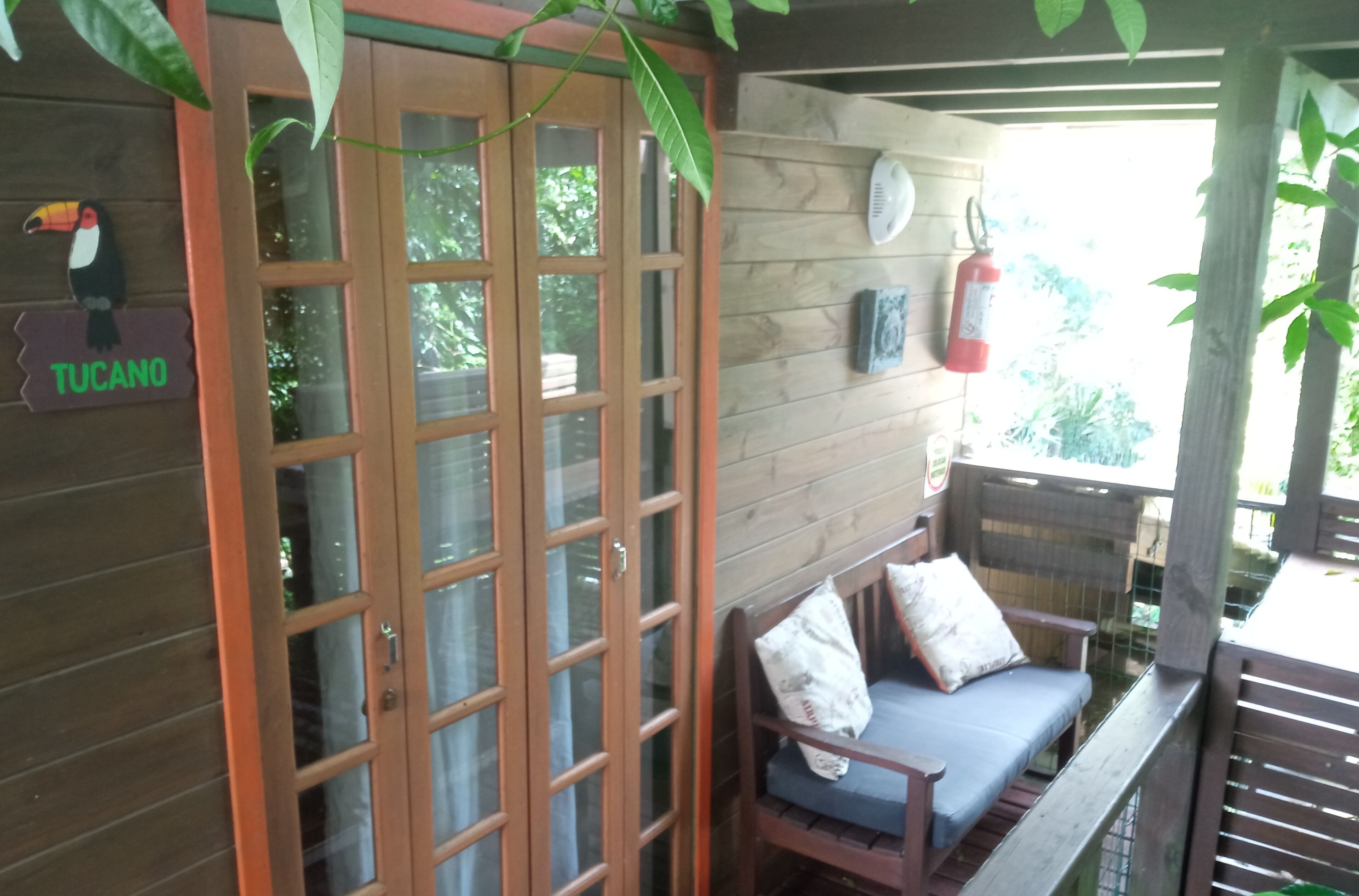 A wooden cabin with a glass door, a bench with cushions, and a sign reading "Tucano".