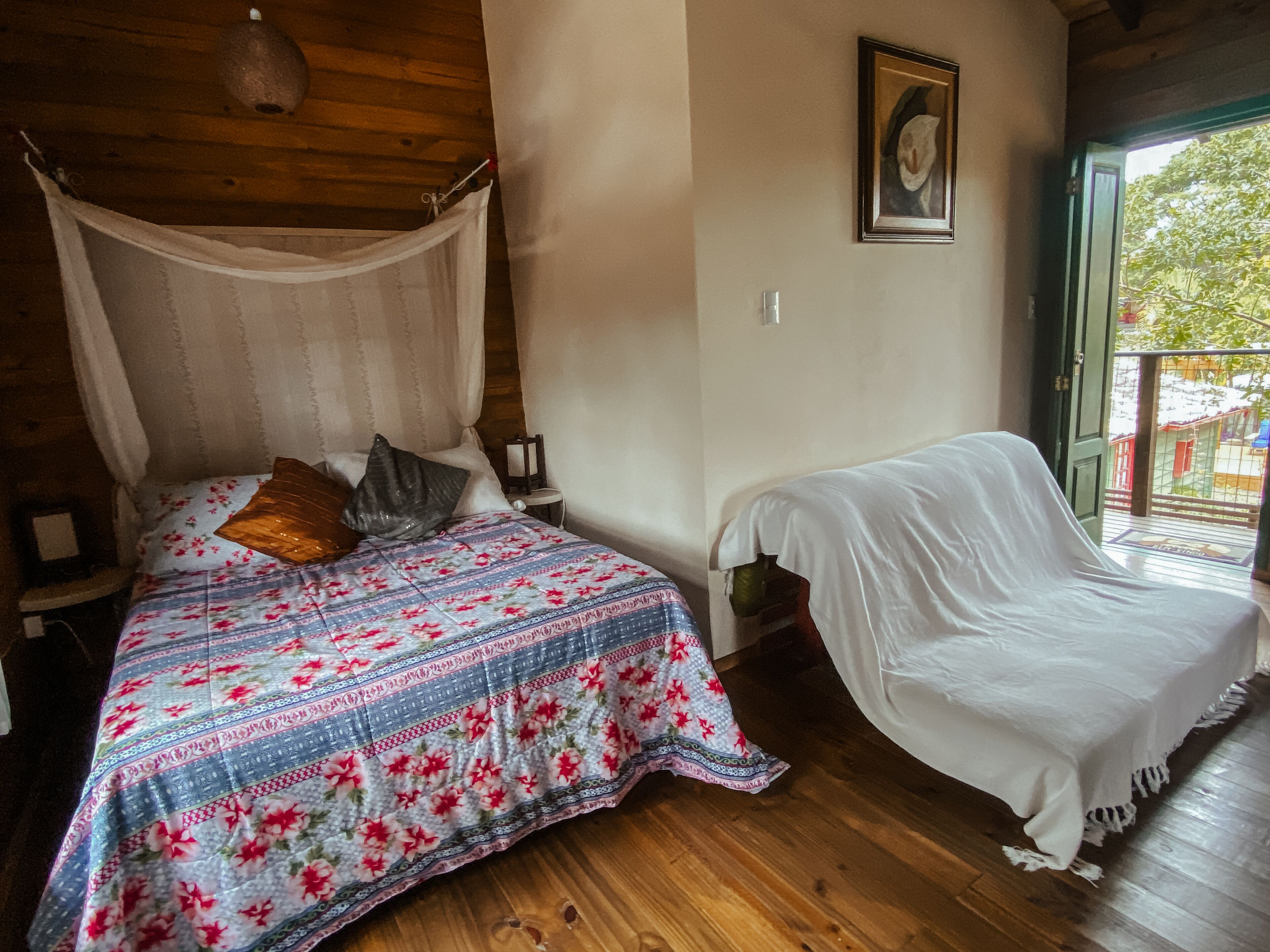 A bedroom with a canopy bed, a bedside table, a chair, and a balcony with a view.