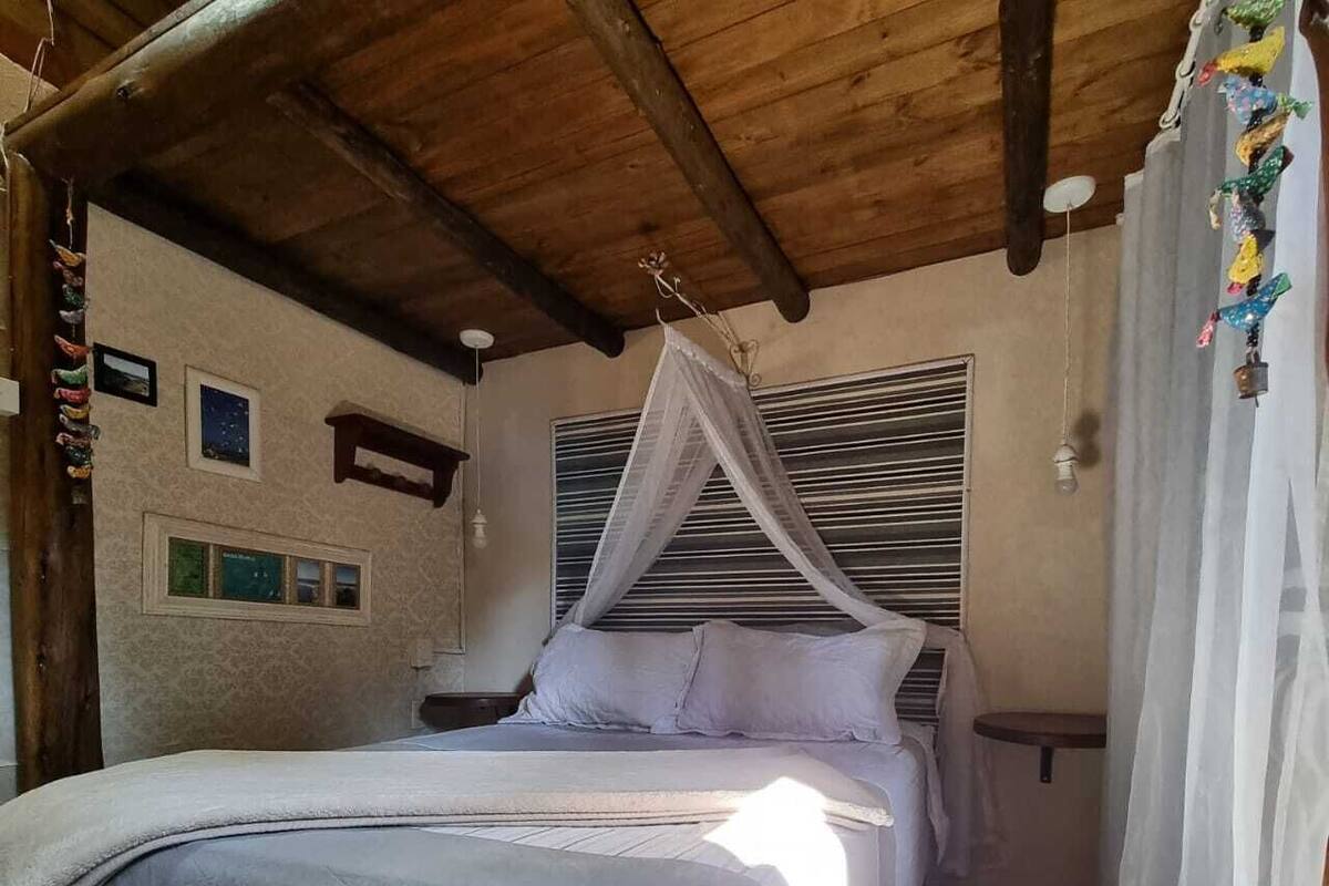 Ein Schlafzimmer mit Bett, Baldachin, Holzbalkendecke, einem kleinen Tisch und einer Wand mit gerahmten Bildern.
