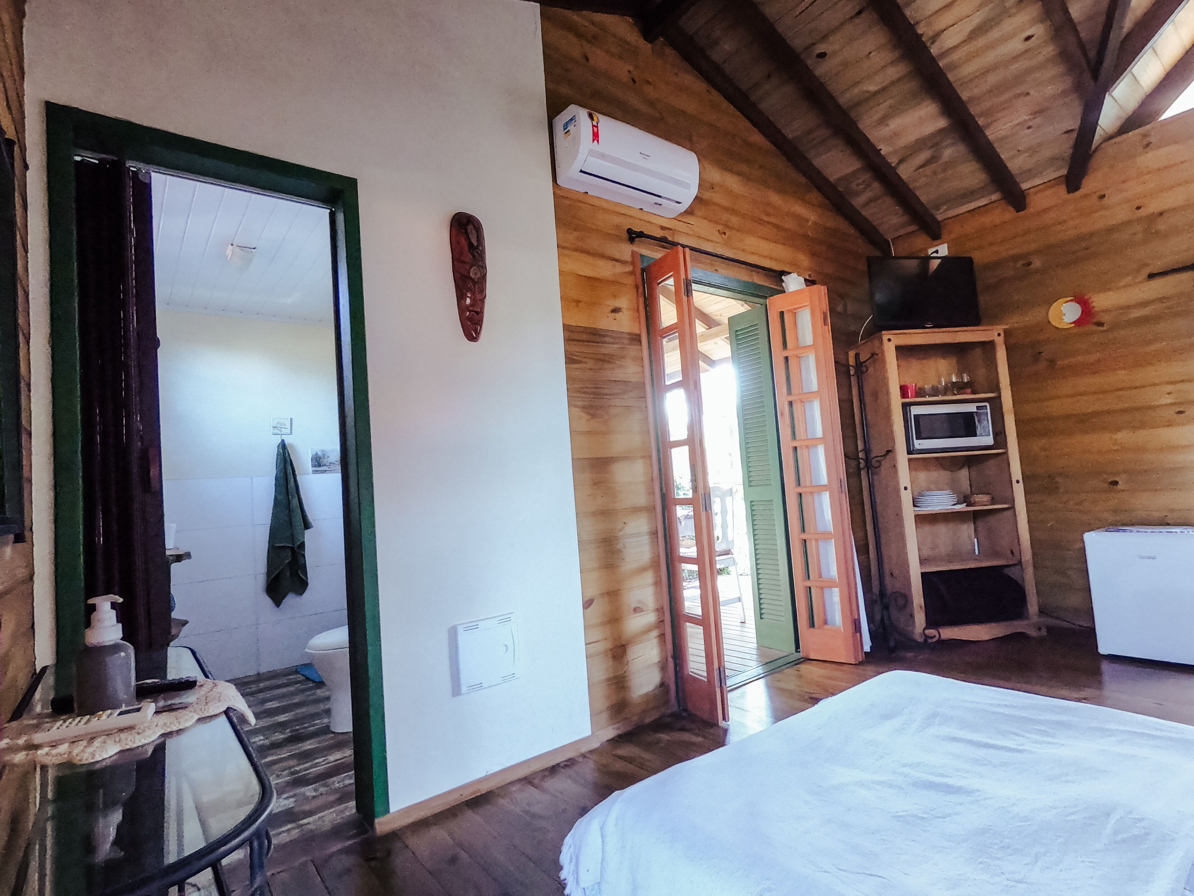 A wooden room with a bed, a door leading to another room, a small table with a bottle and scissors, and a shelf with a microwave and dishes.