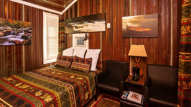 A room with a bed, two chairs, a nightstand with a lamp, and artwork on the walls. - Yowie Cabin (Benton)