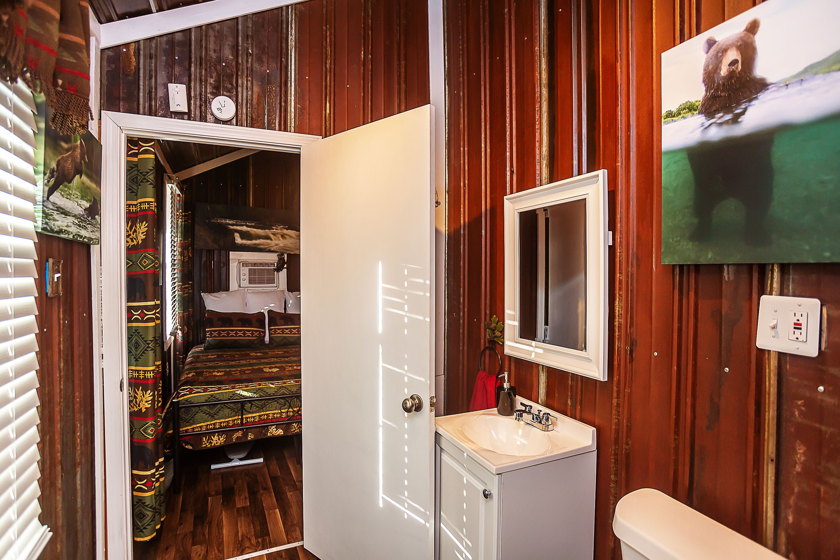 Ein Zimmer mit einer Holzwand, einem Bett mit gemusterten Betttextilien, einem weißen Schrank mit Spiegel und einem Bärenbild an der Wand.