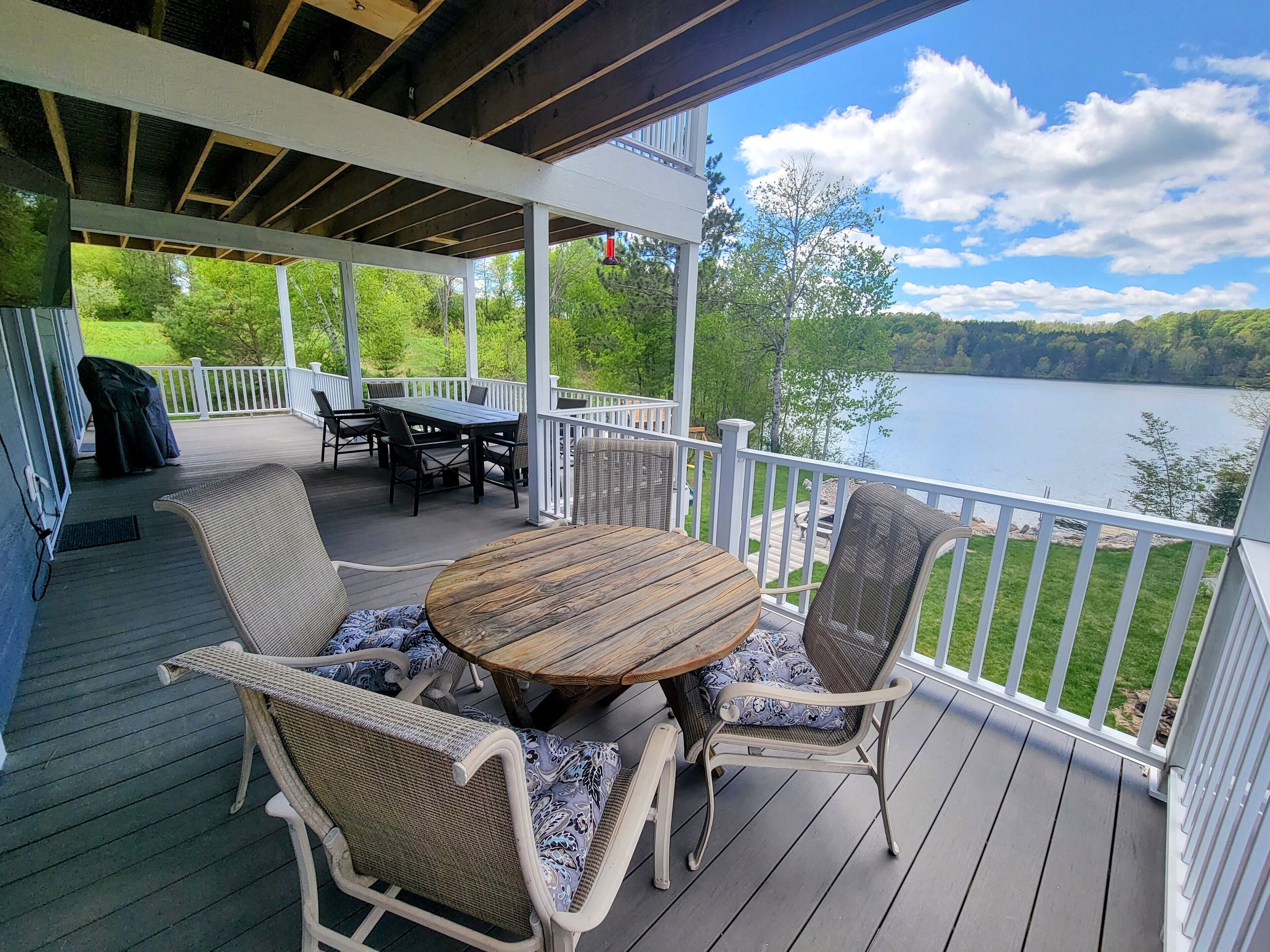 A covered deck with a wooden table and wicker chairs, overlooking a lake.