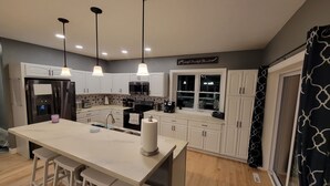 Una cocina moderna con armarios blancos, una isla central con taburetes altos, electrodomésticos de acero inoxidable, iluminación colgante y un ventanal con vistas.