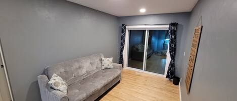 A modern living room with a grey sofa, wooden flooring, and a sliding glass door leading to a balcony.