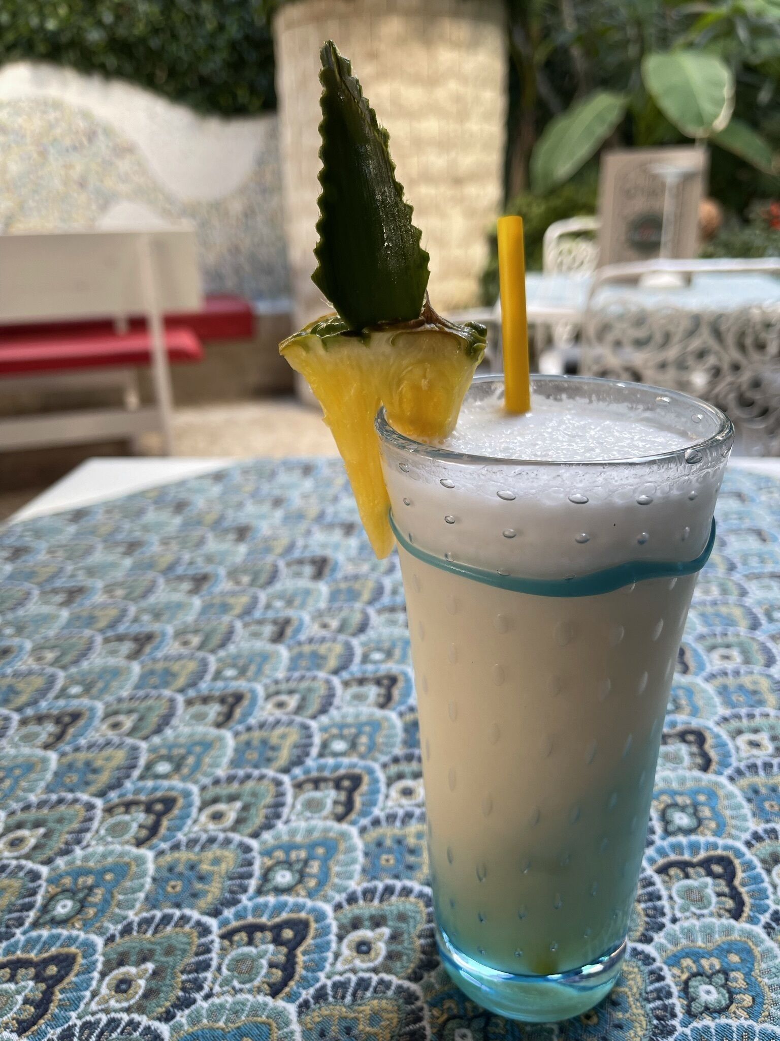 Ein Glas mit schaumigem Getränk, einer Ananas Scheibe und einem Strohhalm auf einer gemusterten Tischdecke.