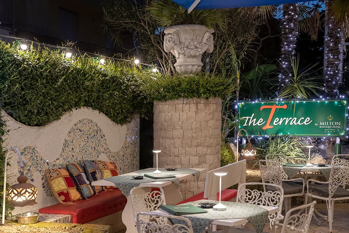 Un área de estar al aire libre con mesas y sillas, un banco acolchado y una pared decorativa. Un letrero dice "The Terrace by Milton Boutique Hotel".