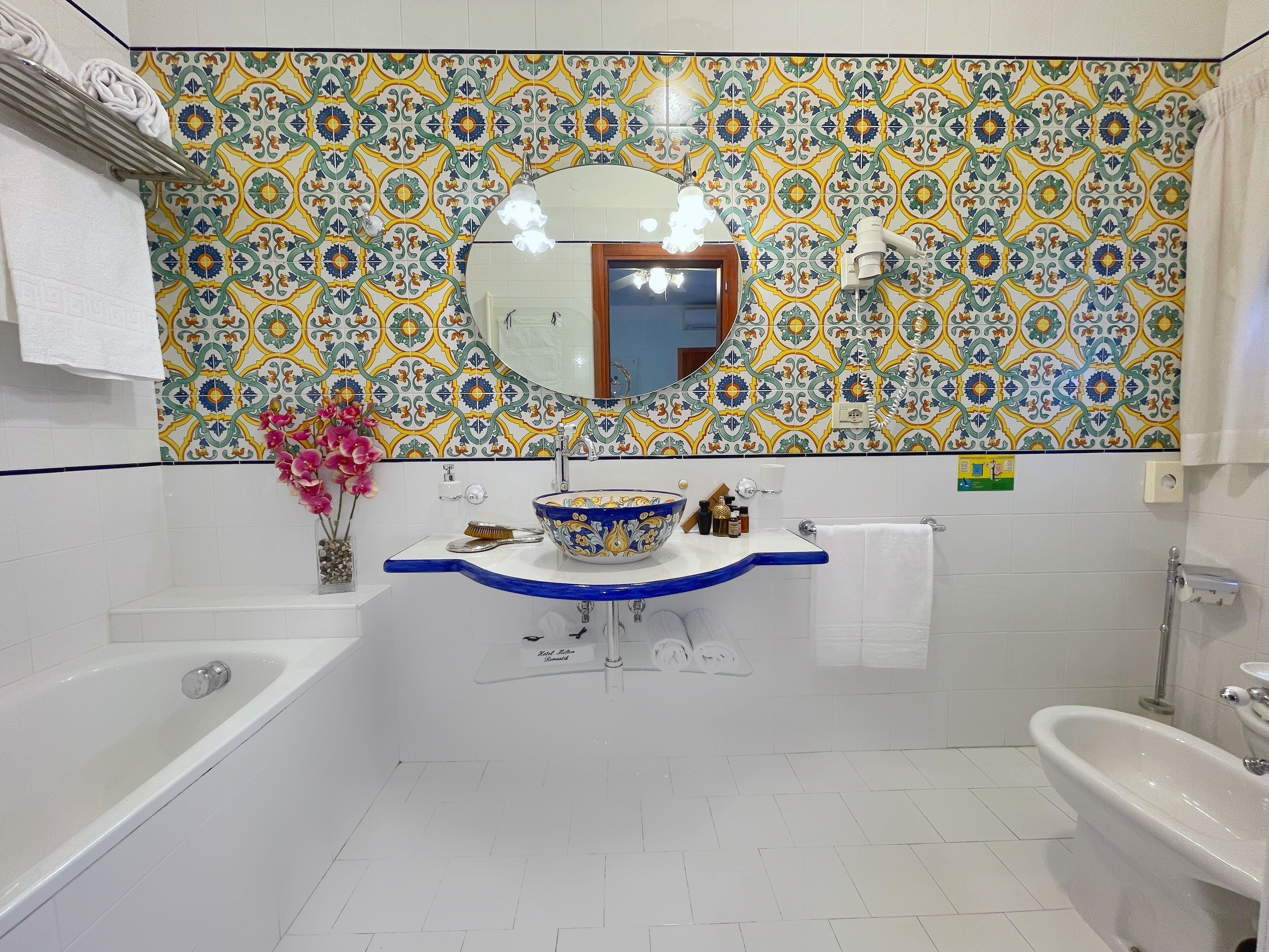 A bathroom with a white bathtub, a sink with a blue basin, and a toilet. The walls are tiled with a colorful pattern.
