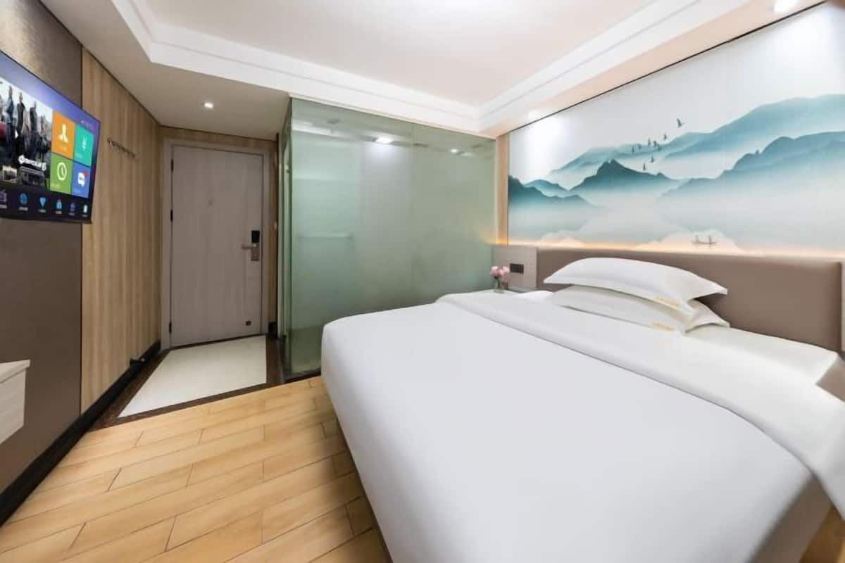 A modern hotel room with a large bed, a glass door leading to a bathroom, and a wall-mounted TV displaying hotel amenities.