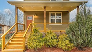 A two-story house with a wooden porch, a staircase, and a well-maintained garden with shrubs and a large coniferous tree.