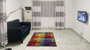 Une pièce avec un canapé bleu foncé, un tapis coloré, un lampadaire, un téléviseur fixé au mur et une fenêtre ornée de rideaux à motifs.