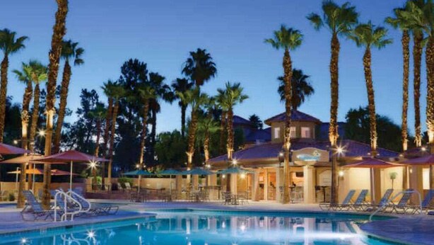 A pool area with lounge chairs and umbrellas, surrounded by palm trees and a building with a red roof. - Marriott Desert Springs II, Luxury 1 bedroom villa (Palm Desert)