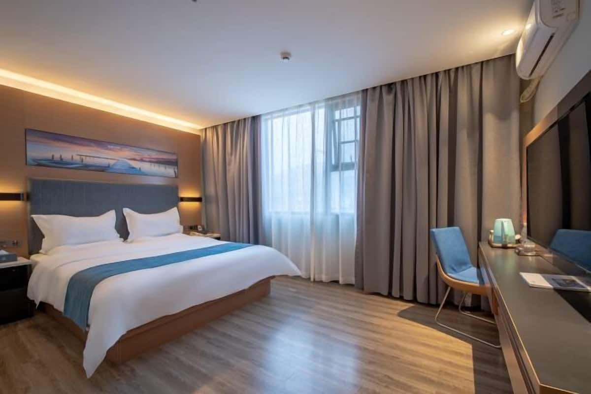 A hotel room with a large bed, a desk with a chair, a painting on the wall, and a window with curtains.