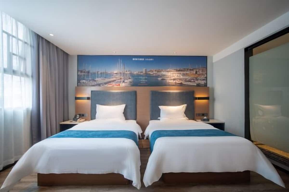 A hotel room with two beds, a cityscape mural on the wall, and a large window with curtains.