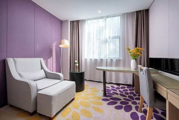 Una moderna camera d'hotel con un divano grigio, un piccolo tavolino rotondo, una televisione e un'ampia finestra con tende.