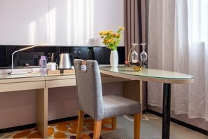 Una moderna camera d'albergo con una scrivania in vetro, una poltrona grigia rivestita in tessuto, un vaso con fiori gialli, un portabicchieri per vino e una TV a schermo piatto.