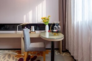 Una camera d'albergo con una scrivania, una sedia, un tavolino rotondo con un vaso di fiori, una bottiglia di vino e una lampada.