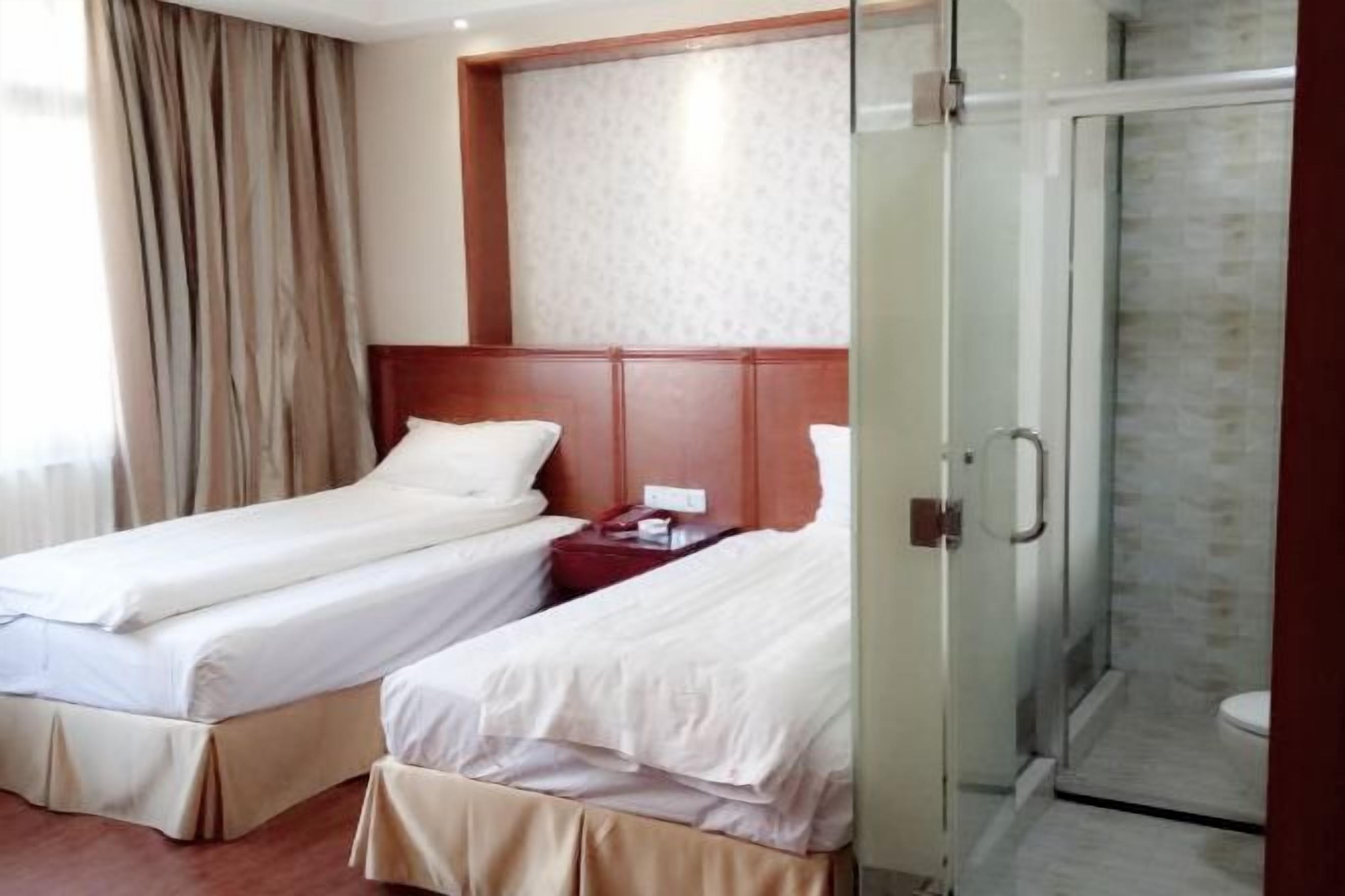 Quarto de hotel com duas camas de solteiro, cabeceira de madeira, box de vidro para o chuveiro e parede com padrão.