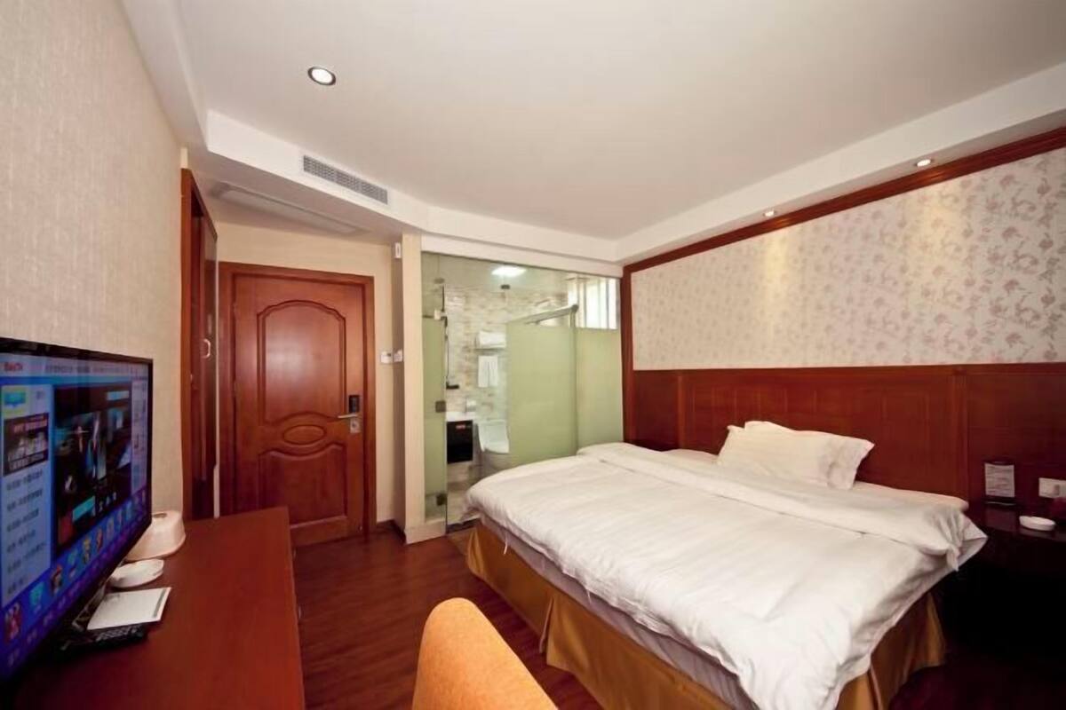 A hotel room with a bed, a desk with a computer, and a bathroom visible through a glass door.