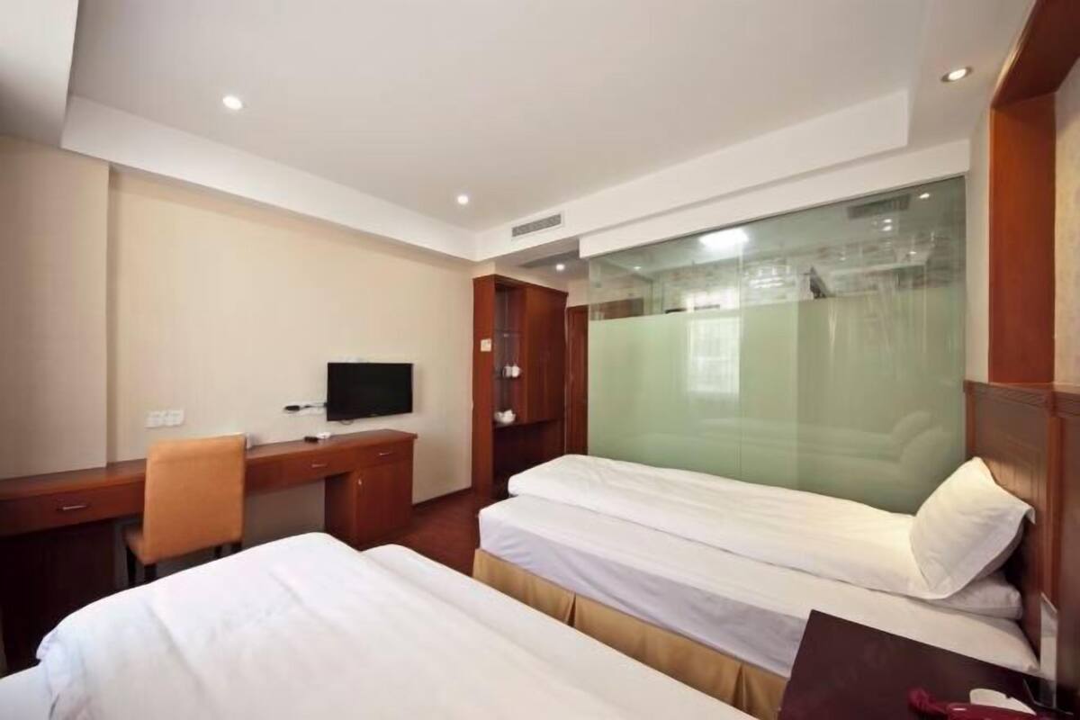 A hotel room with two beds, a desk, a chair, a television, and a glass partition.