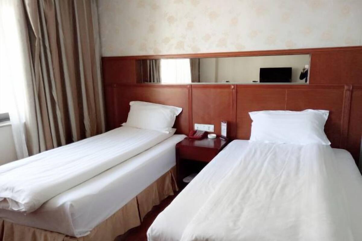 A hotel room with two single beds, a wooden headboard, a bedside table with a lamp, and a mirror above the headboard.
