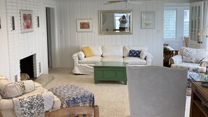 Een ruime woonkamer met een open haard, een groene salontafel en diverse zitoogjes, waaronder een bank en fauteuils.