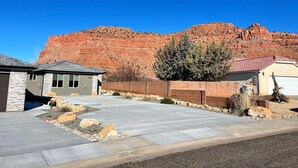 A residential area with a paved driveway, a stone building, and a large red rock formation in the background.