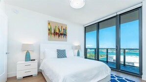 Une chambre moderne avec un grand lit, deux tables de chevet avec des lampes, un tableau coloré représentant un paysage urbain, et un balcon avec vue sur la mer.