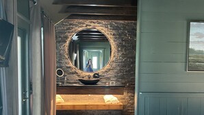 A hallway with a wooden floor, a stone wall, a large mirror, a sink, and a bed.