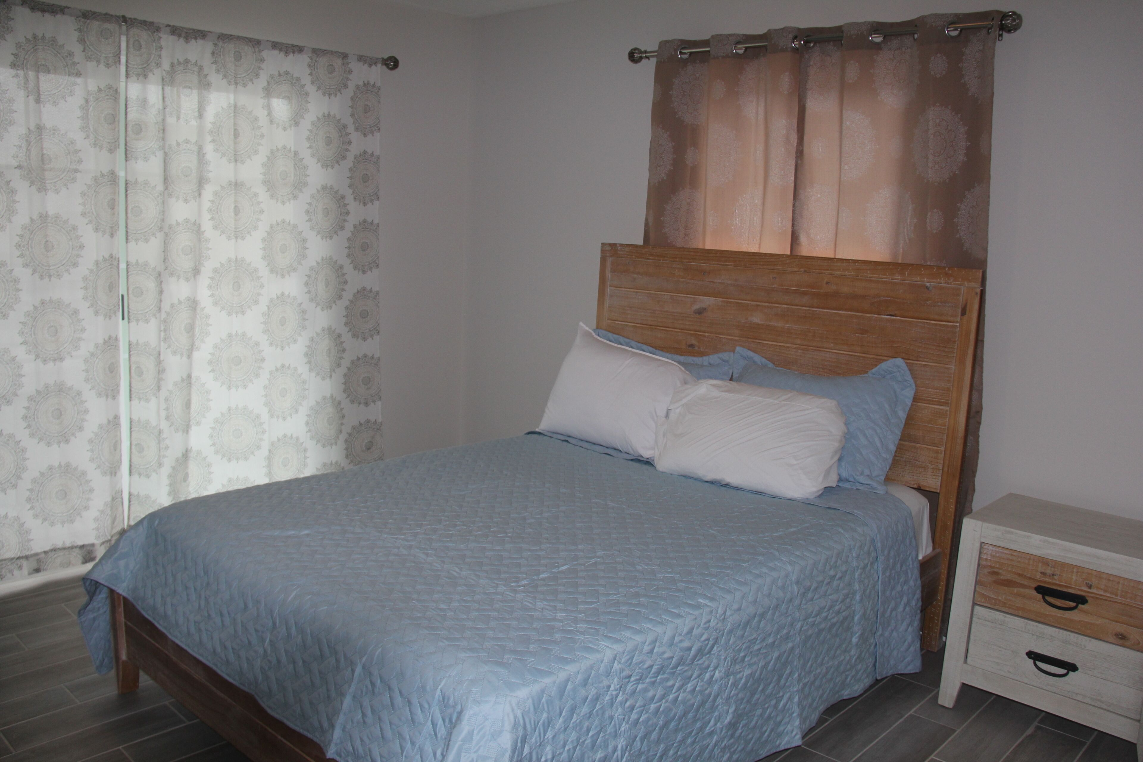 A bedroom with a wooden bed frame, a blue bedspread, two pillows, a nightstand with drawers, and a curtain with a floral pattern.