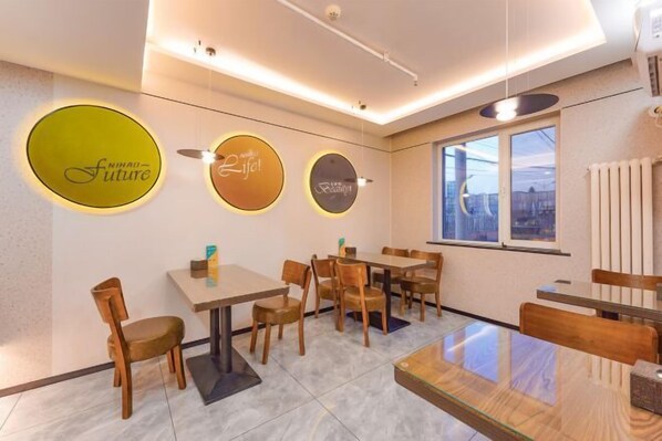 Interior de restaurante con mesas y sillas redondas, vista a la ciudad a través de amplios ventanales y letreros iluminados en la pared.