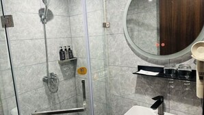 A bathroom with a glass-enclosed shower, a round mirror, a white sink, and a shelf with toiletries.